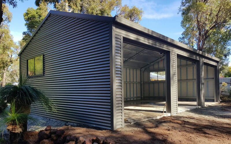 Exterior side view of a large, dark grey corrugated steel shed with three open roller bays surrounded by native trees