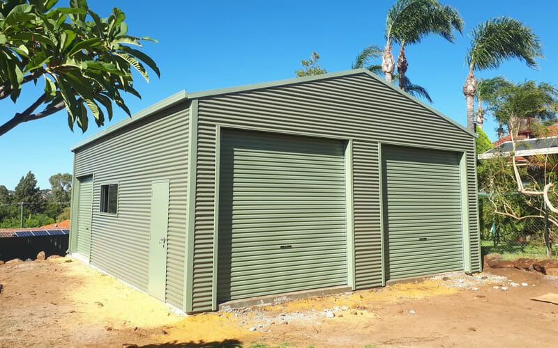 Exterior of a new pale green steel shed with two closed roller doors and a side door, built on a residential block