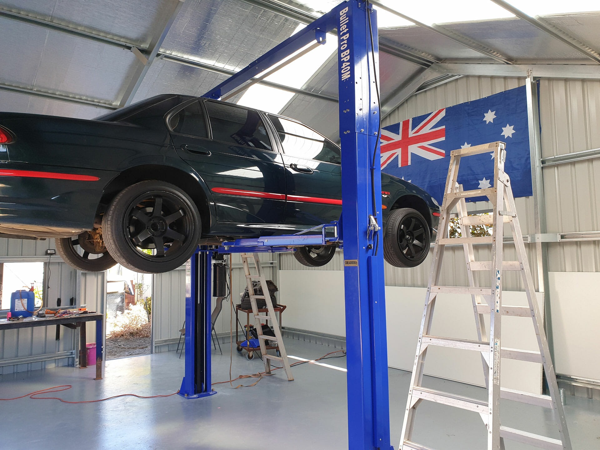 A black car lifted on a blue two-post hoist inside a spacious metal workshop, maximising the shed's vertical space